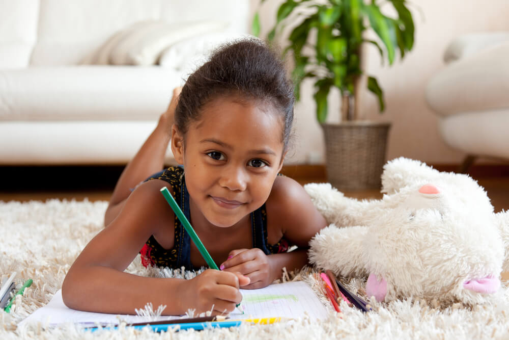 girl laying on the carpet floor in a living room drawing on paper with markers