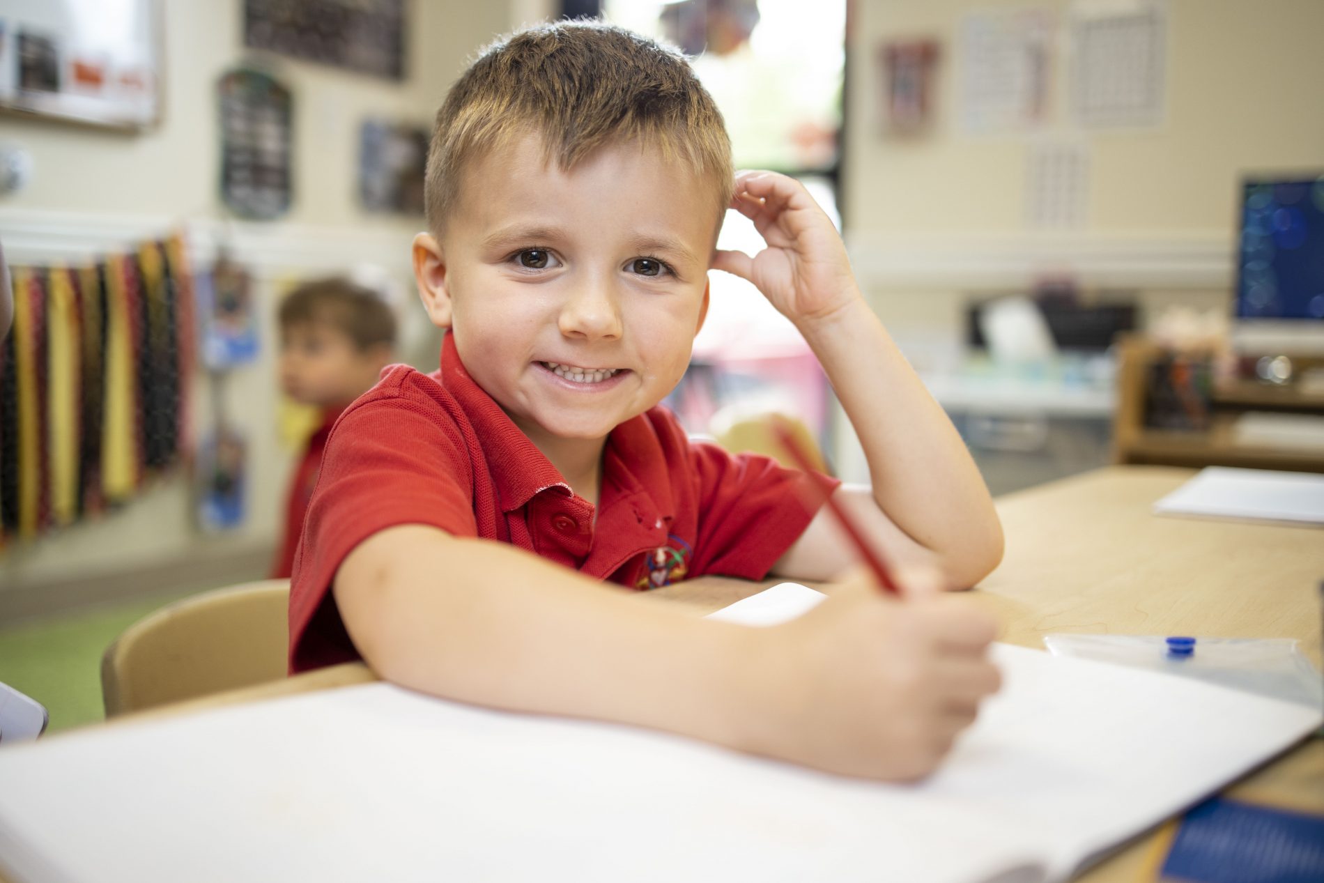 young boy smiling while holding a pencil sitting at a school desk in a red polo uniform