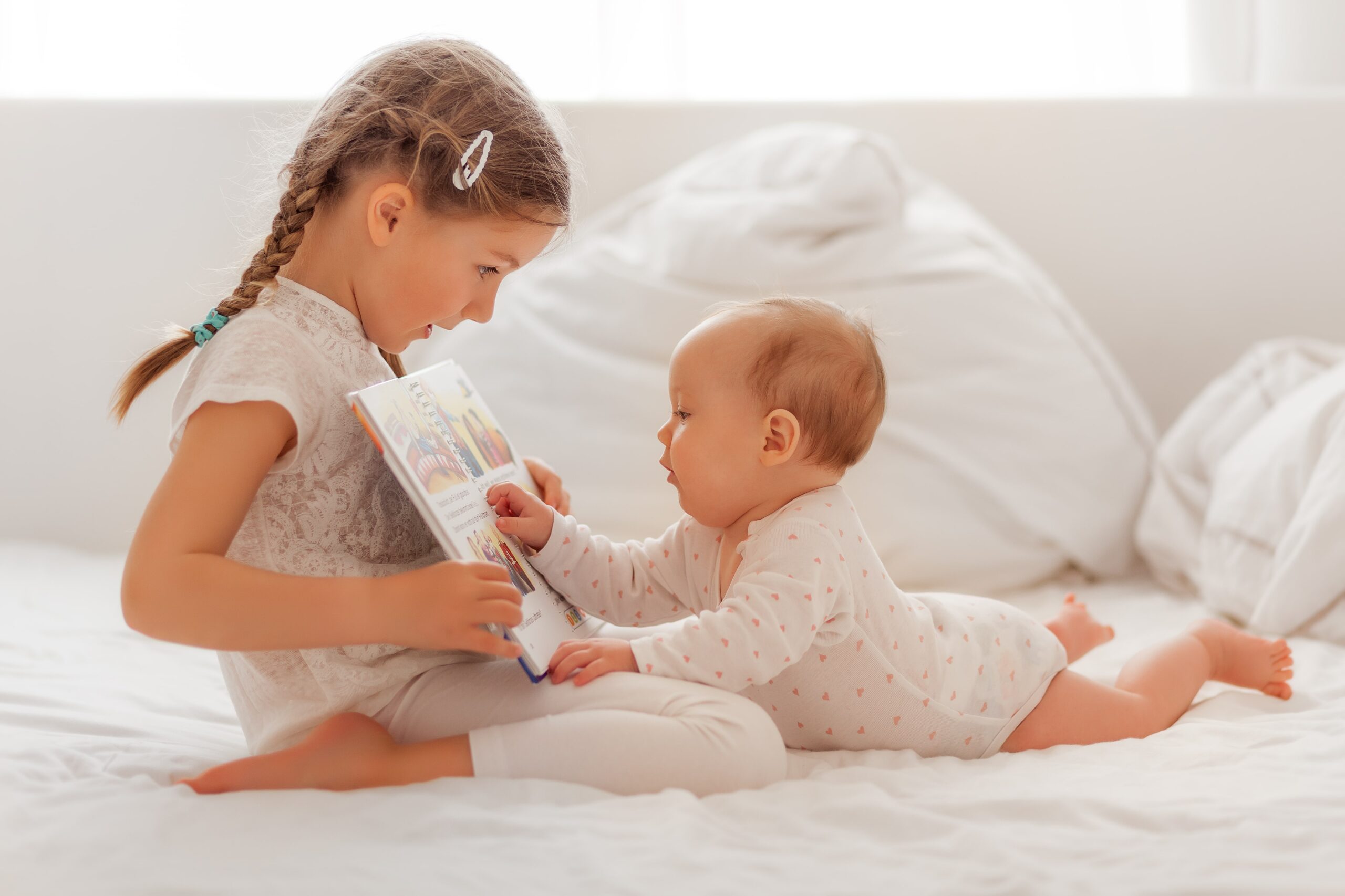 young girl reading book to baby sibling on bed